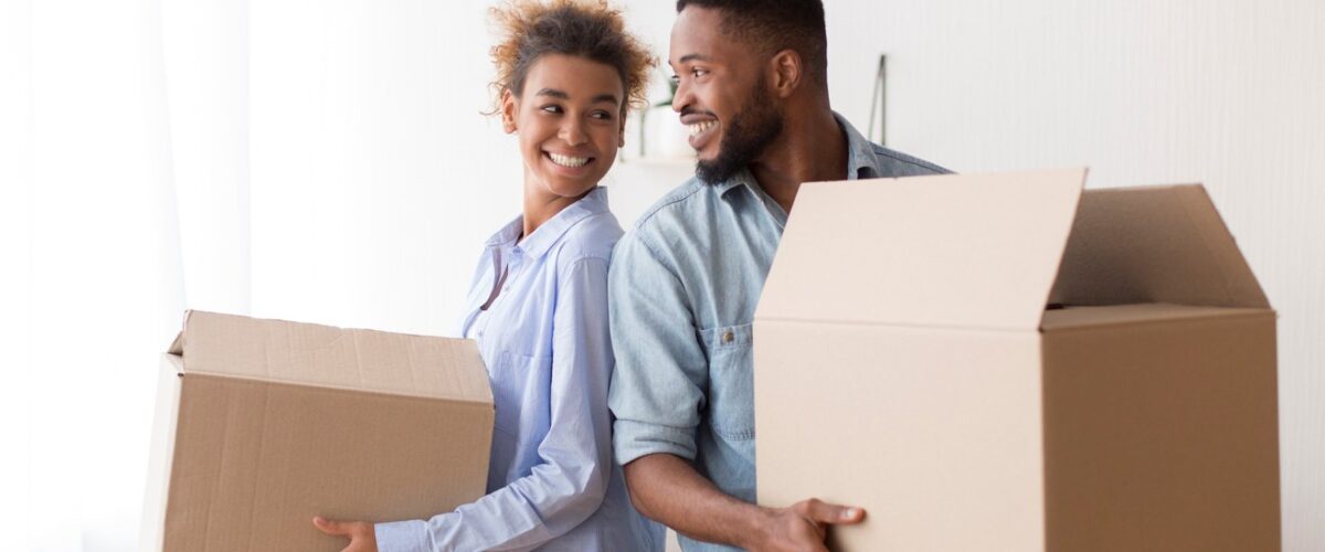 A young couple standing back-to-back smile at each other while holding cardboard boxes.