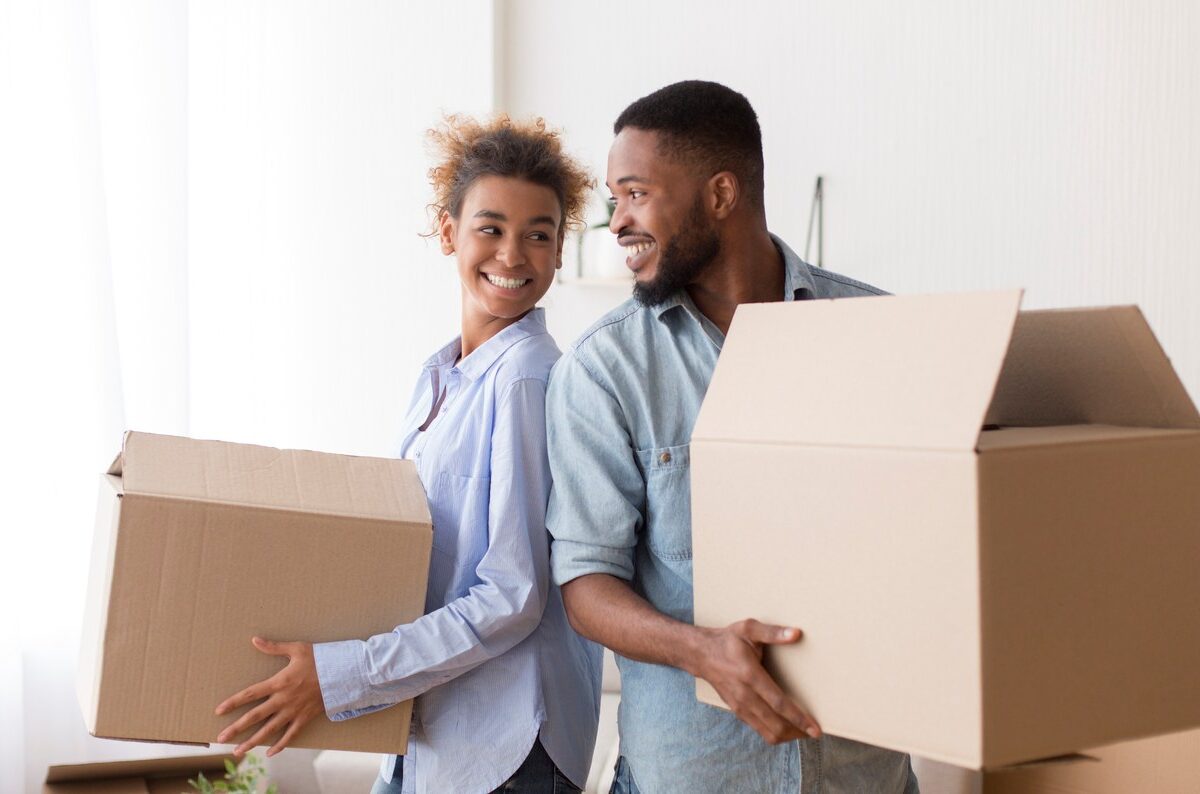 A young couple standing back-to-back smile at each other while holding cardboard boxes.