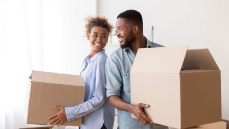 A young couple standing back-to-back smile at each other while holding cardboard boxes.