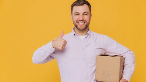 A man in a button-up shirt holds a box and gives a thumbs-up.