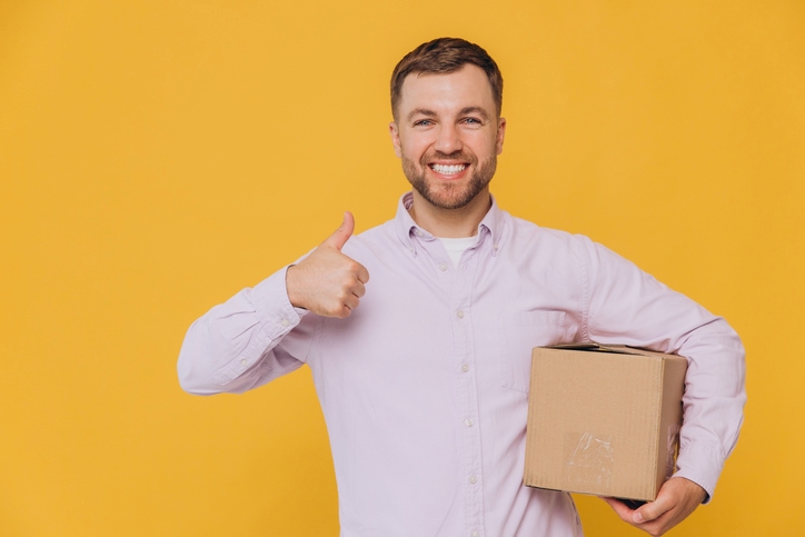 A man in a button-up shirt holds a box and gives a thumbs-up.