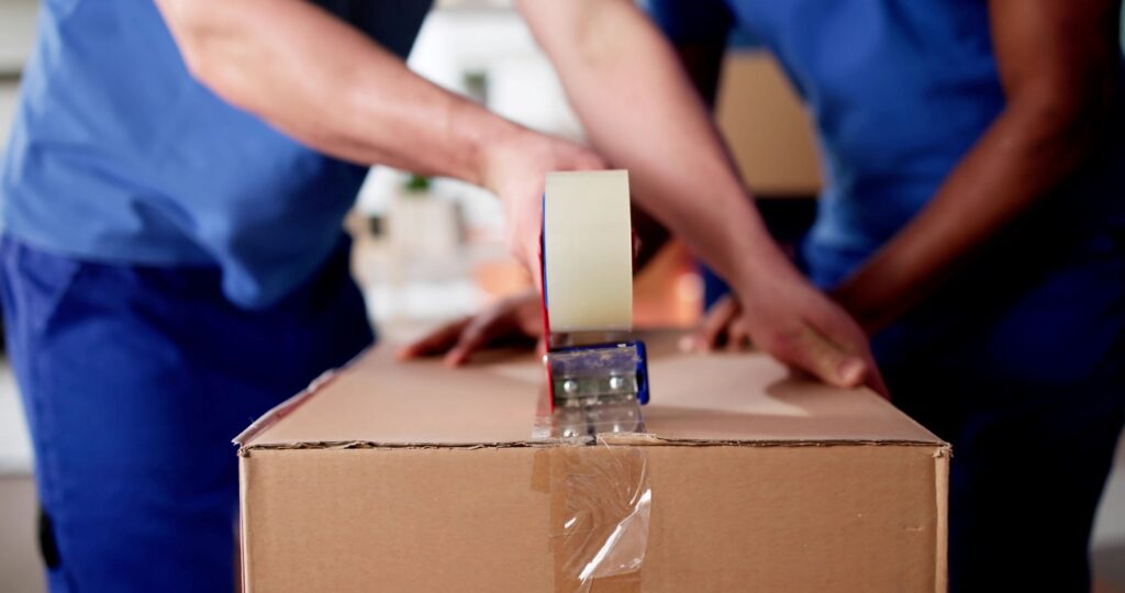 Two movers in blue uniforms use packing tape to seal off a cardboard box.