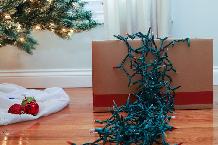 Holiday string lights spilling from a cardboard box sit under a holiday tree, next to red and green glass ornaments.